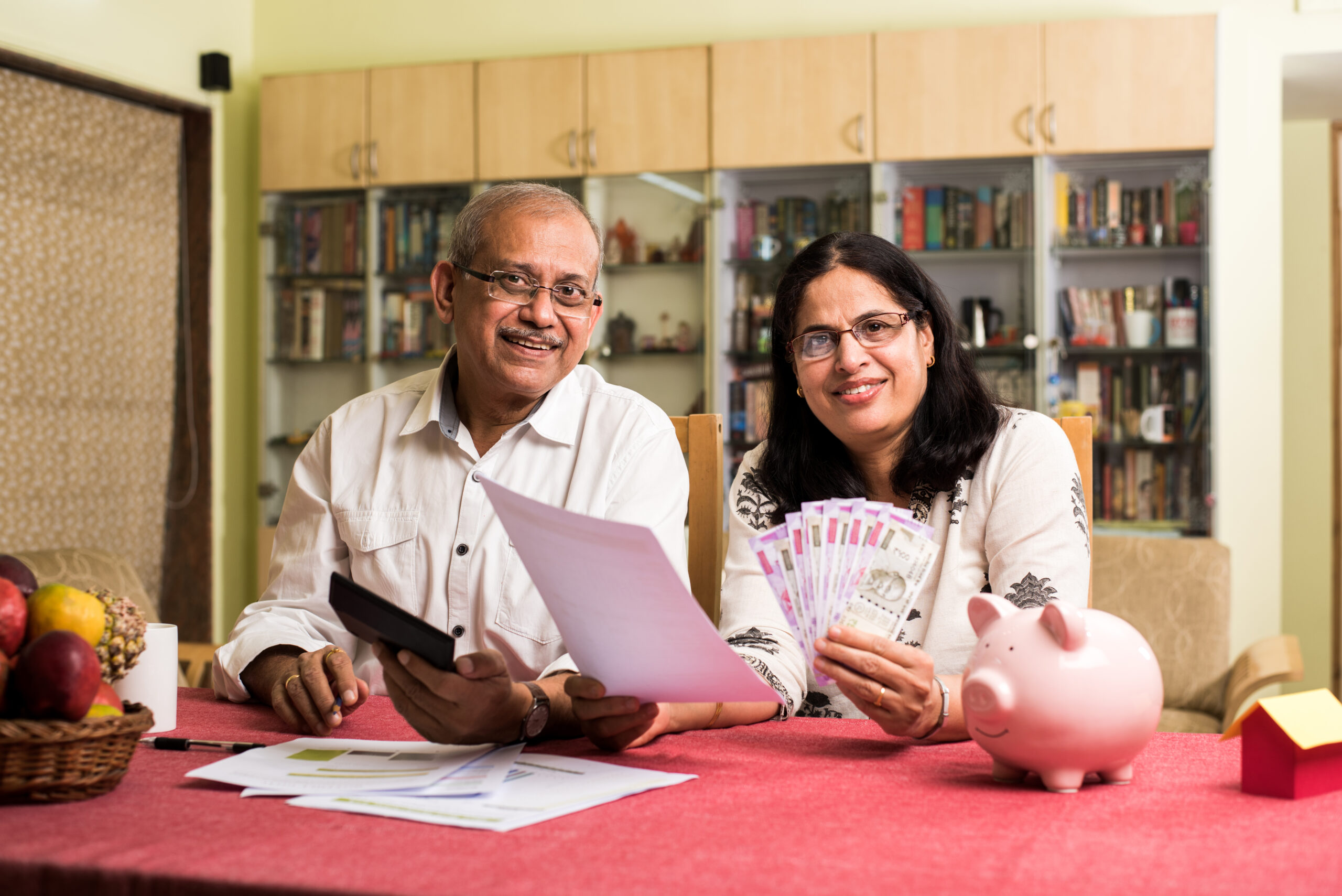 Senior Indian asian couple accounting, doing home finance and checking bills with laptop, calculator and money also with piggy bank while sitting on sofa couch or table at home